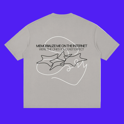 A light gray short-sleeve t-shirt with a black graphic print on the back. The graphic features the text 'MEMORIZE ME ON THE INTERNET' and 'WERE THE ONES YOU LEAST EXPECT' above a line-art illustration of three overlapping star-like shapes.